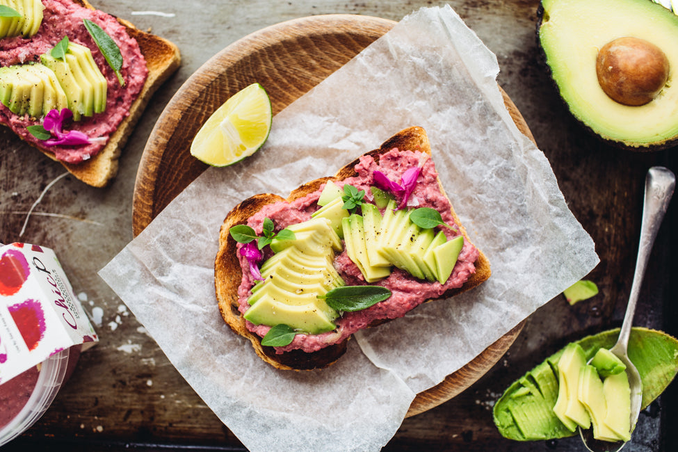 ChicP's bright beetroot hummus on a piece of toast with slices of avocado and fresh herbs. A slice of lime and the other half of the avocado is placed next to it 